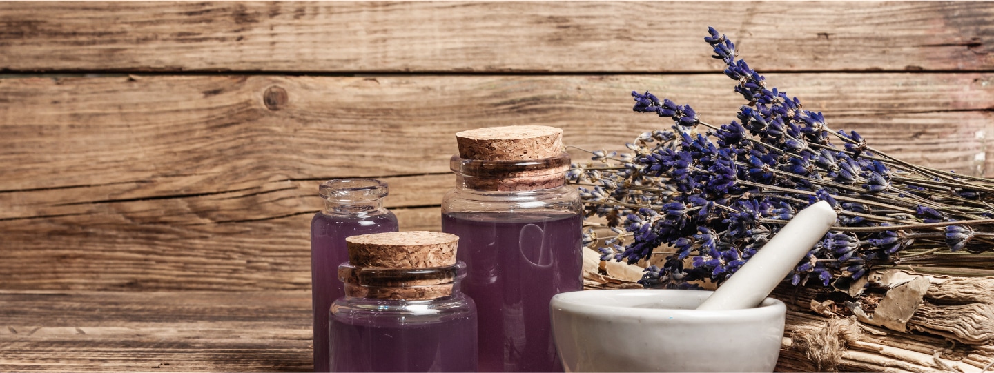 Drei Glasfläschchen mit lila Flüssigkeit, ein Mörser mit Stößel und getrockneter Lavendel auf Holztisch vor Holzbretter-Hintergrund.